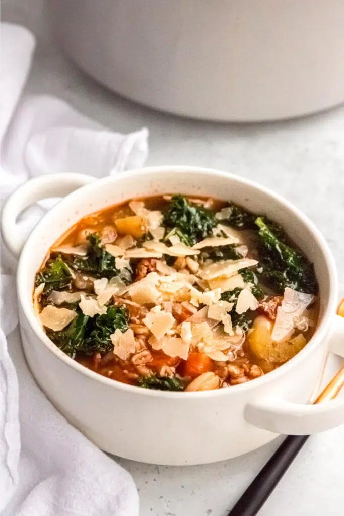 Rustic Tuscan soup with beans, grains, and greens, topped with cheese in a white bowl.
