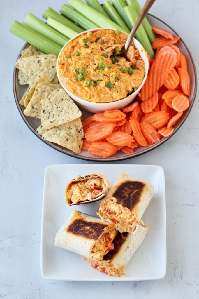 A close-up of a halved buffalo chicken wrap with a tangy, cheese-filled center, alongside a serving bowl of buffalo dip and raw veggies