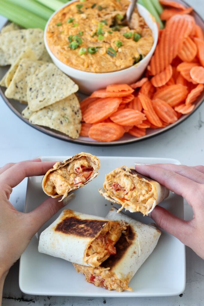 Hands holding a buffalo chicken wraps cut in half to show the creamy filling, with a bowl of buffalo dip surrounded by chips and carrot sticks in the background.