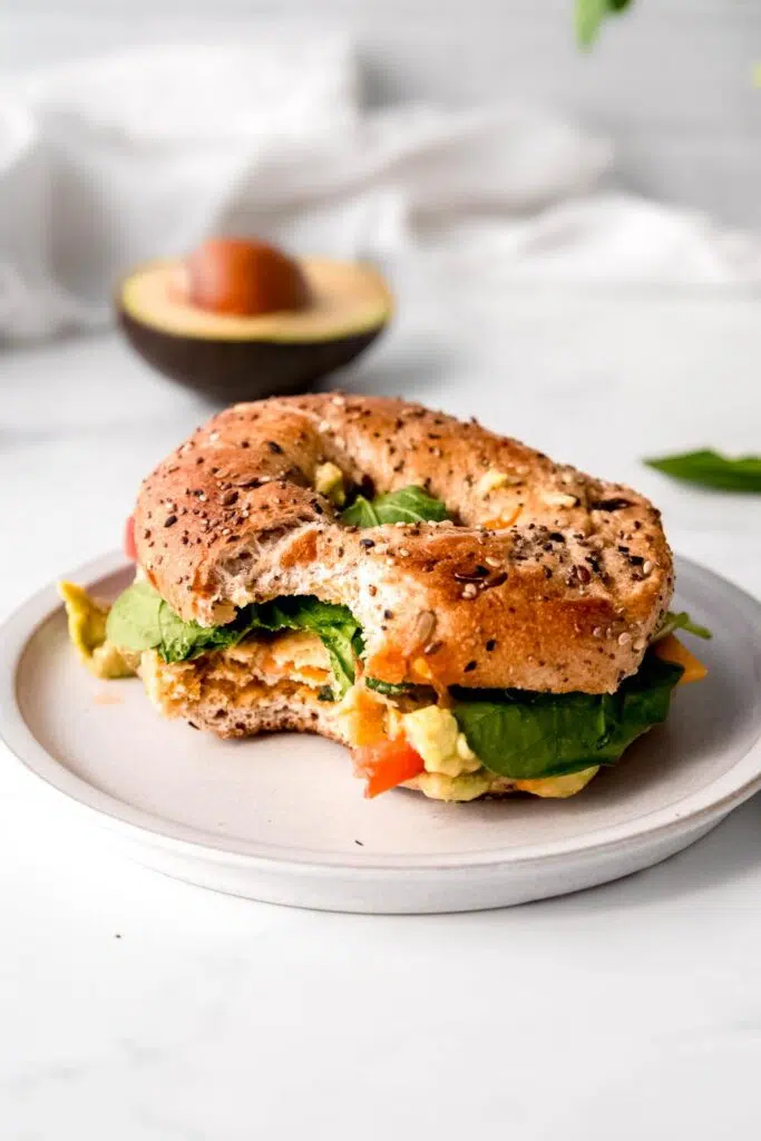 A wholesome bagel sandwich with a bite taken on a ceramic plate, featuring a hearty egg, cheese, spinach, and tomato filling.