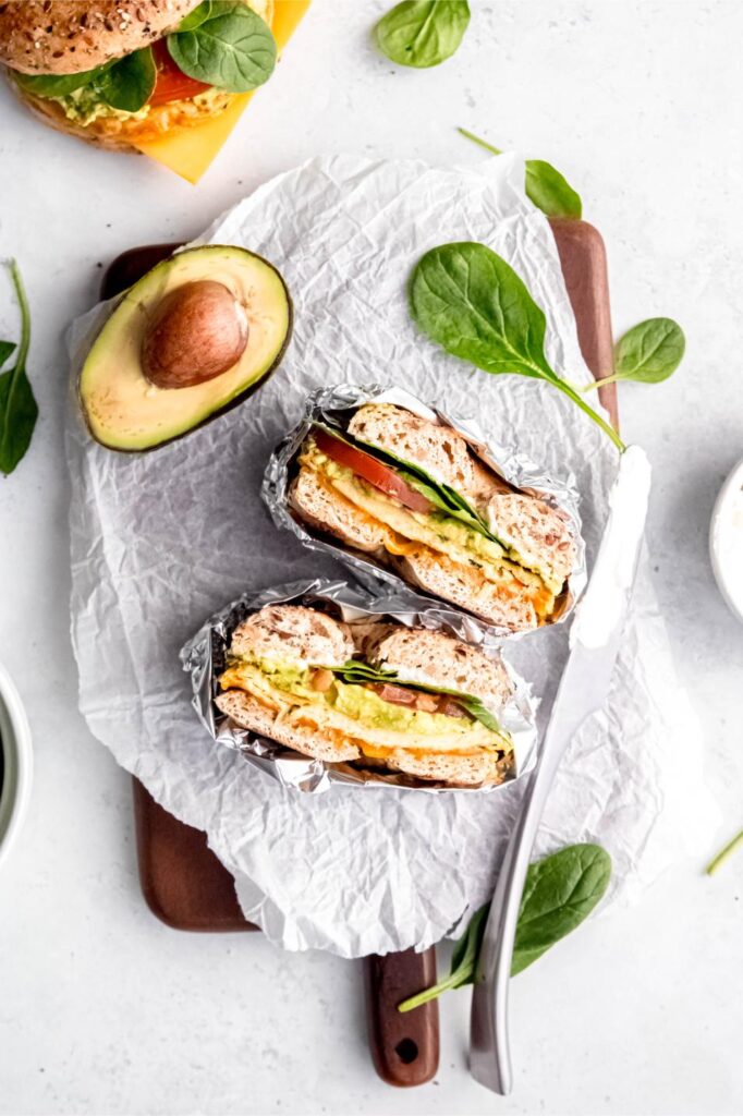 A breakfast setup with a halved, foil-wrapped bagel sandwich, next to a ripe, sliced avocado and a sprinkle of spinach leaves, on a rustic chopping board.