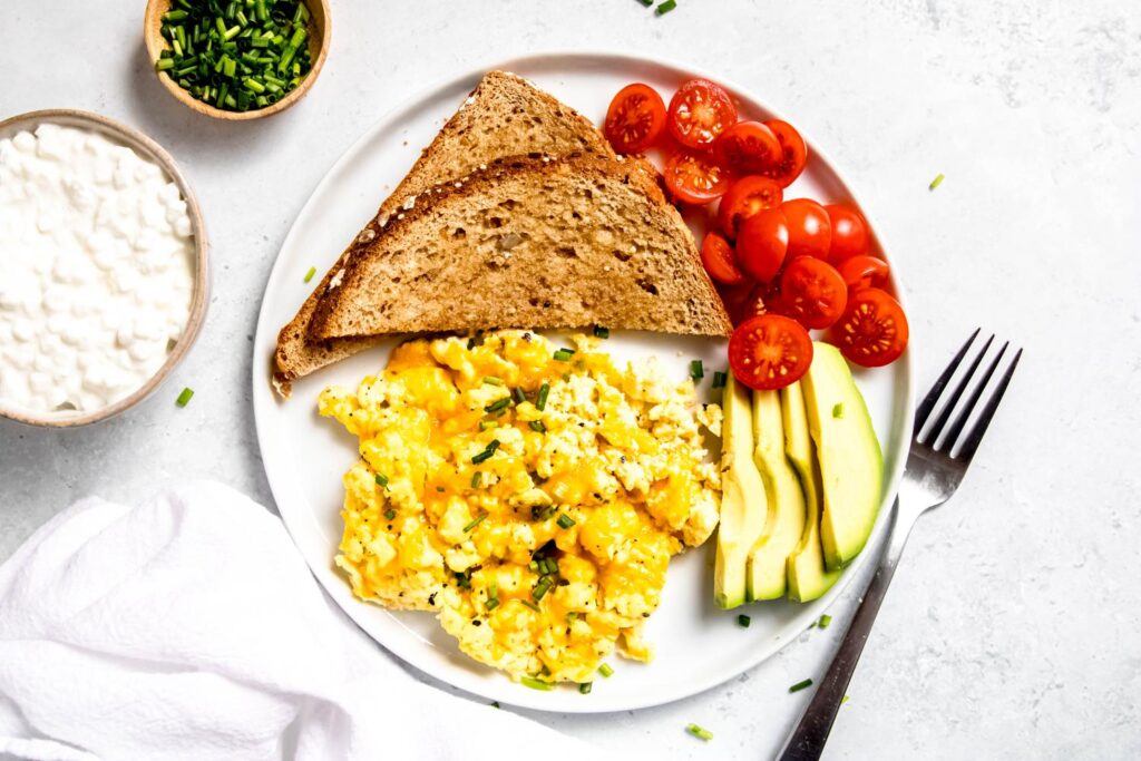 White plate with scrambled cottage cheese eggs with sliced avocado, sliced tomatoes, and sliced whole wheat bread