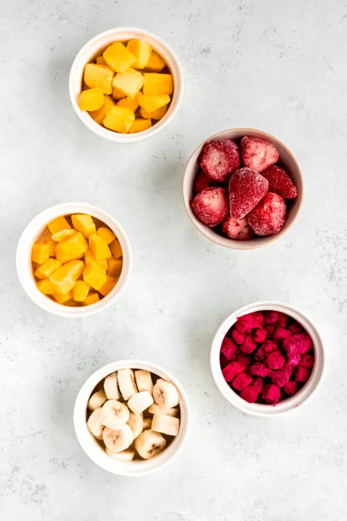 Frozen fruits like strawberries, mango. pineapple, banana, and dragonfruit in white bowls.