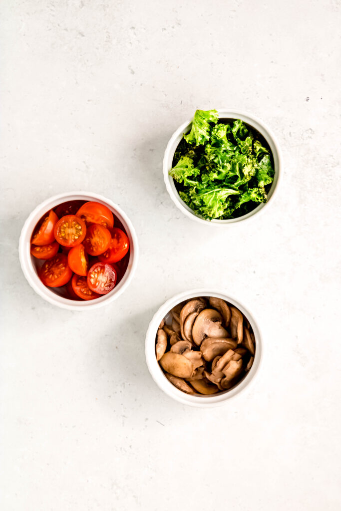 3 white bowls, one with kale, one with sliced tomatoes, and the other with sliced mushrooms.