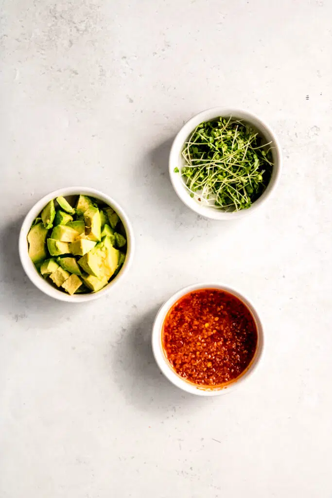 3 white bowls, one with micro greens, one with avocado, and one with chili crisps.