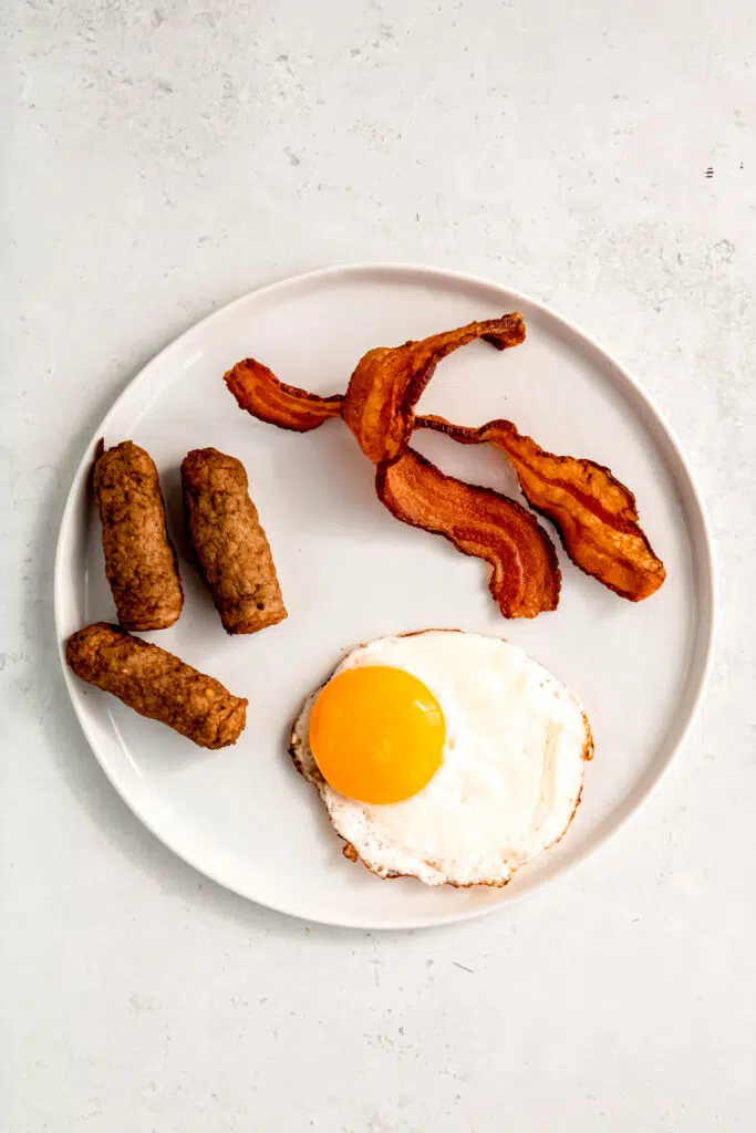 White plate with a sunny side up egg, three sausage lines, and two slices of bacon.