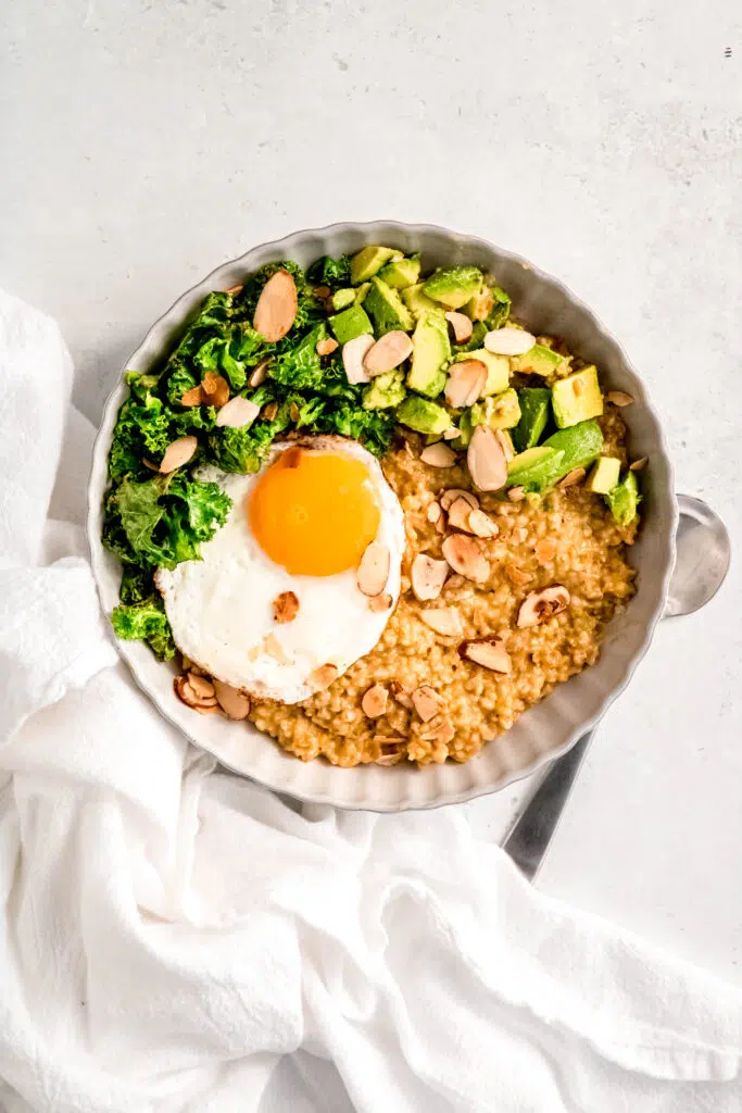 Savory oats with sunny side up egg, greens, avocado, and almonds in white bowl with spoon next to bowl.