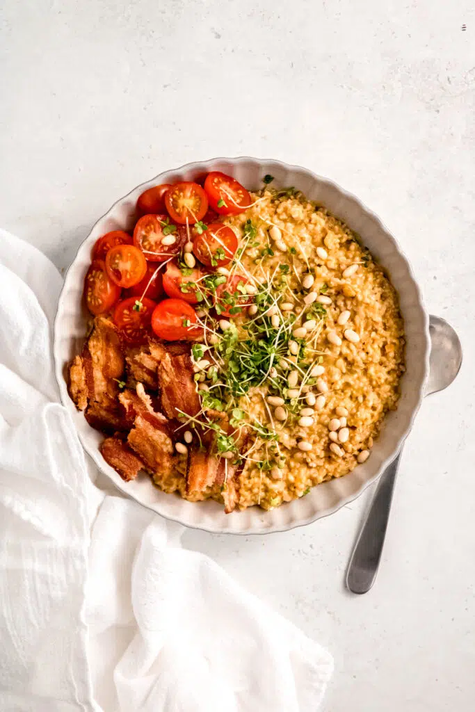 Savory oats with tomatoes, bacon, and pine nuts in white bowl with spoon next to bowl.