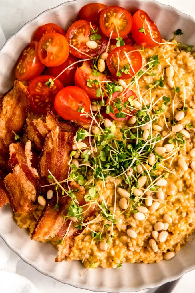 Savory oats with tomatoes, bacon, micro greens, and pine nuts in white bowl. 