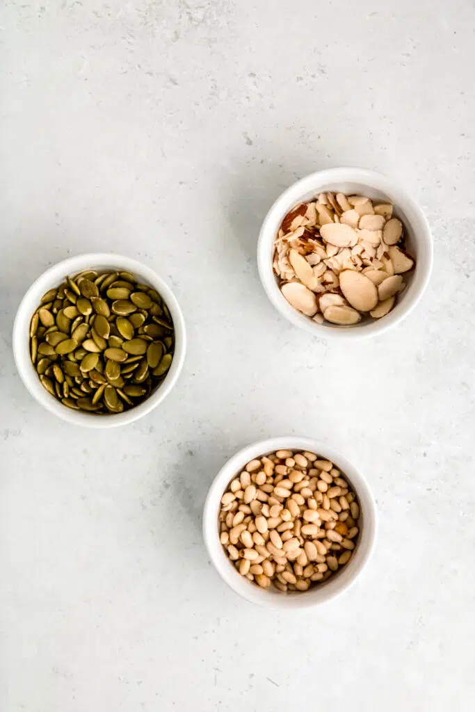 3 white bowls, one with sliced almonds, one with sunflower seeds, and one with pinenuts.