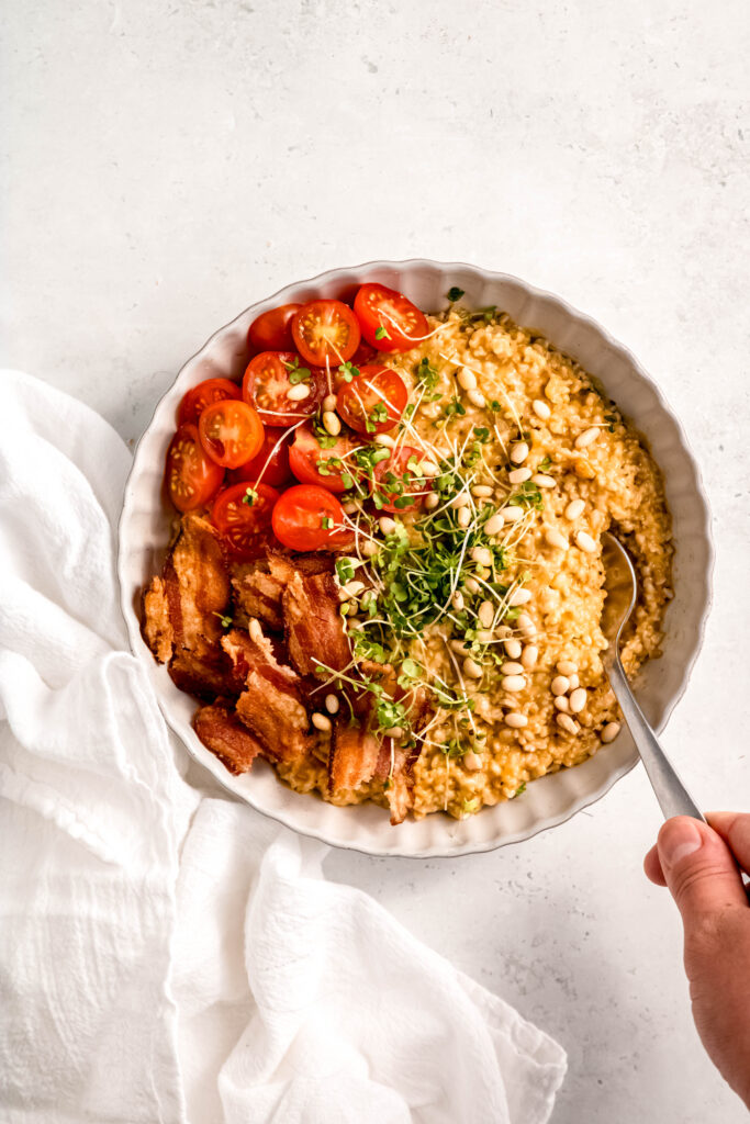 Person holding spoon in savory oats with tomatoes, bacon, micro greens, and pine nuts in white bowl. 