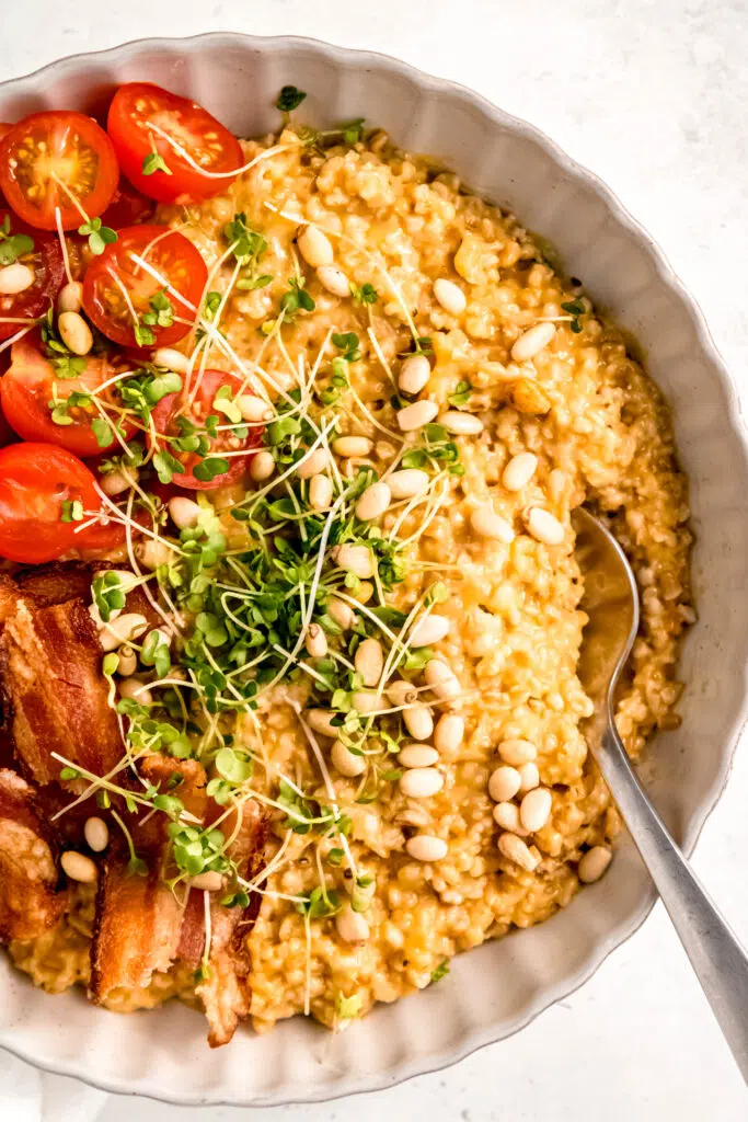 Savory oats recipe template: serves 4 variation with spoon in savory oats with tomatoes, bacon, micro greens, and pine nuts in white bowl 