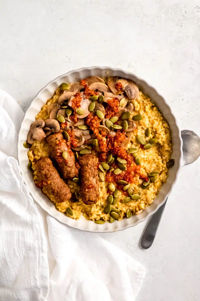 Savory oats with sausage links, mushrooms, and sunflower seeds in white bowl with spoon next to bowl.