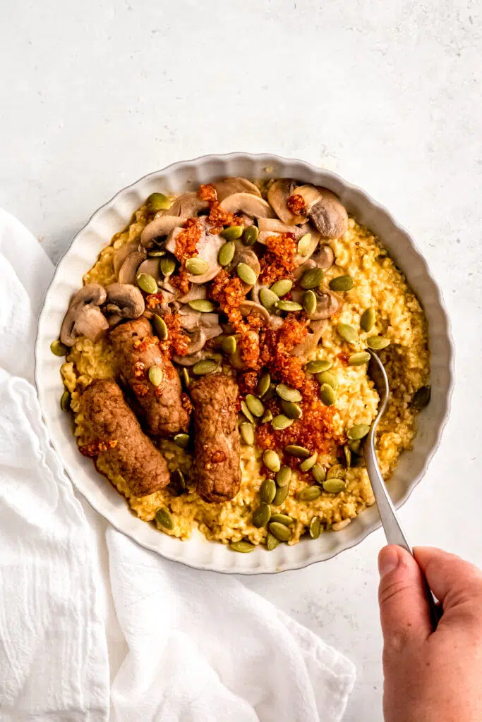 Person holding spoon in savory oats with sausage links, mushrooms, and sunflower seeds in white bowl.