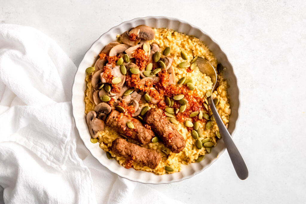 Savory oats with sausage links, mushrooms, and sunflower seeds in white bowl with spoon in bowl.