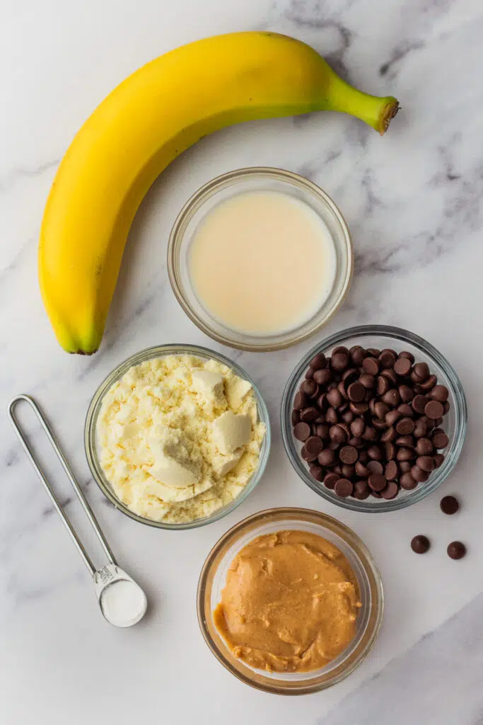 Ingredients for a protein mug cake, including a banana, protein powder, chocolate chips, peanut butter, and milk, arranged on a marble surface. A simple guide to making a protein cake in a mug.