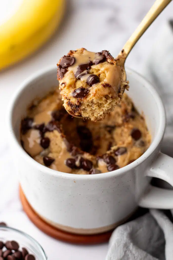 Freshly baked chocolate protein microwave cake in a white mug, with gold spoon lifting it up