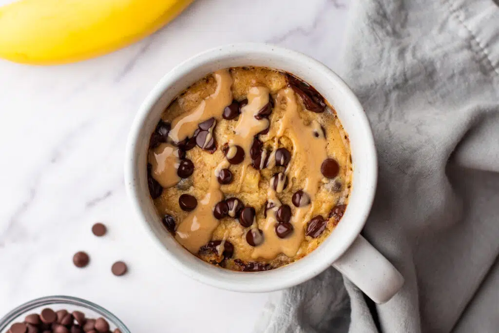 Close-up of a protein powder mug cake topped with chocolate chips and peanut butter, served in a white mug on a marble countertop.