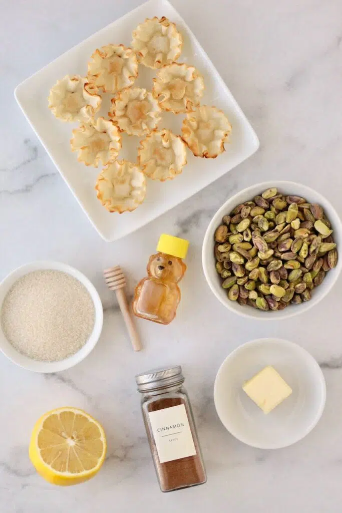 Ingredients laid out: phyllo shells, pistachios in bowl, sugar, honey, butter, lemon, and cinnamon