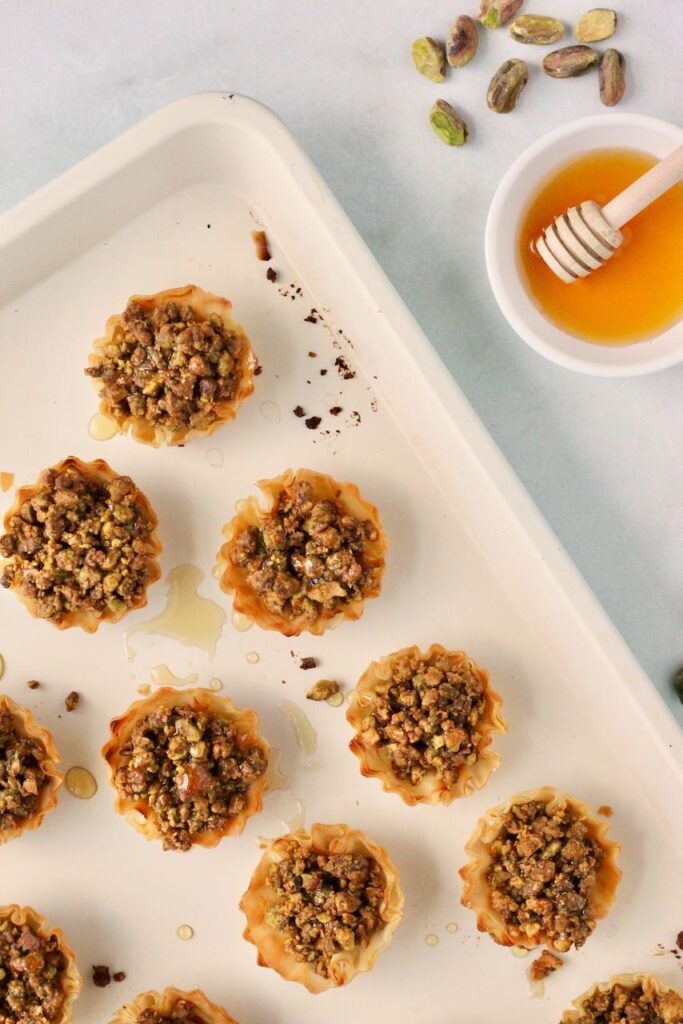 Baklava bites on baking sheet with pistachios and honey around