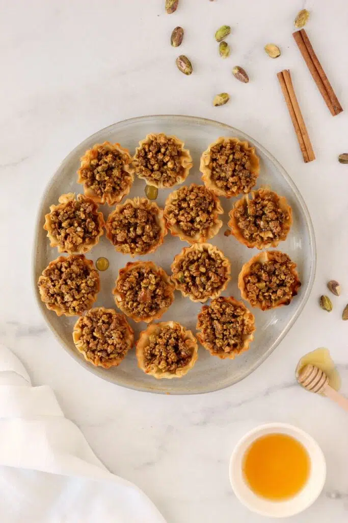 Baklava bites on plate with honey, pistachios, and cinnamon sticks around