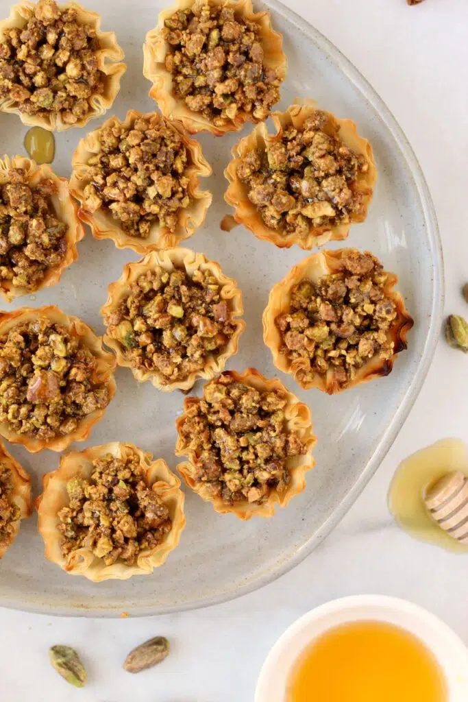 Close up of baklava bites on plate with honey and pistachios around plate