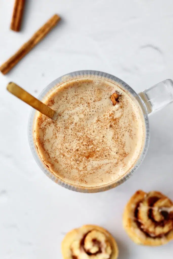 Cinnamon roll protein latte with cinnamon sticks and cinnamon rolls in background