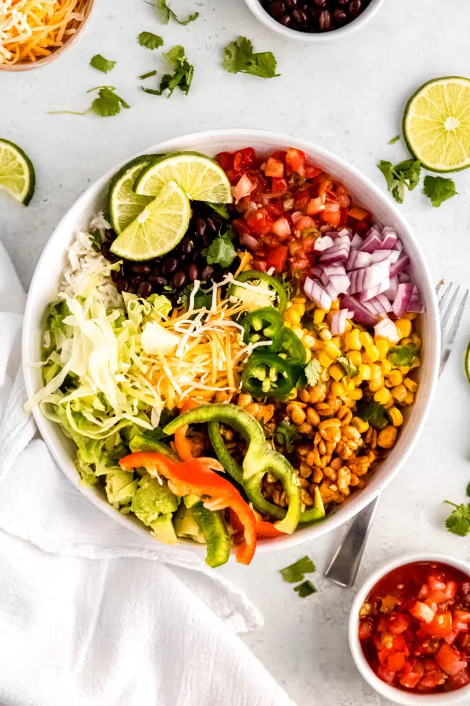 Finished tempeh burrito bowl with sliced peppers, jalapeños, red onion, lettuce and lime wedges. 