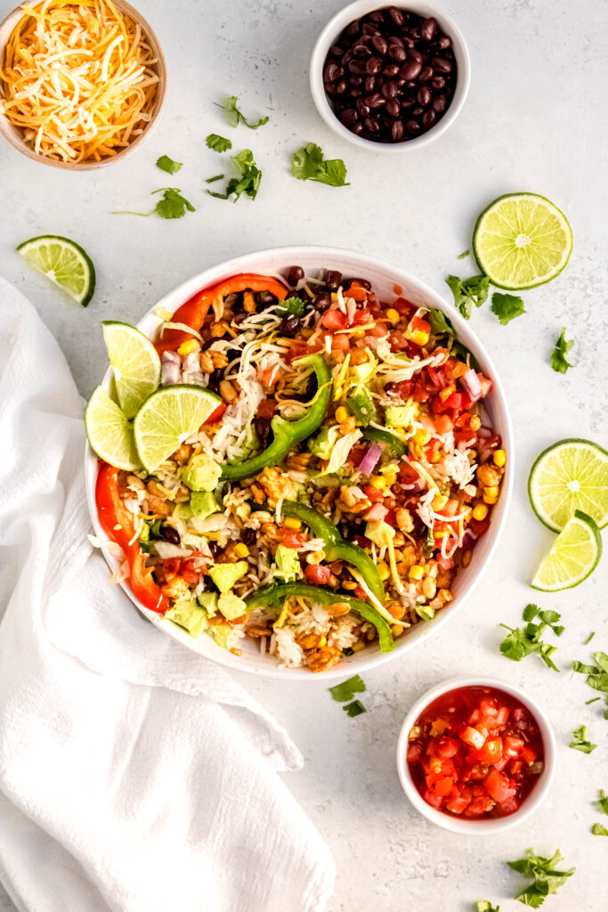 Finished tempeh burrito bowl with sliced peppers, jalapeños, red onion, lettuce and lime wedges. 