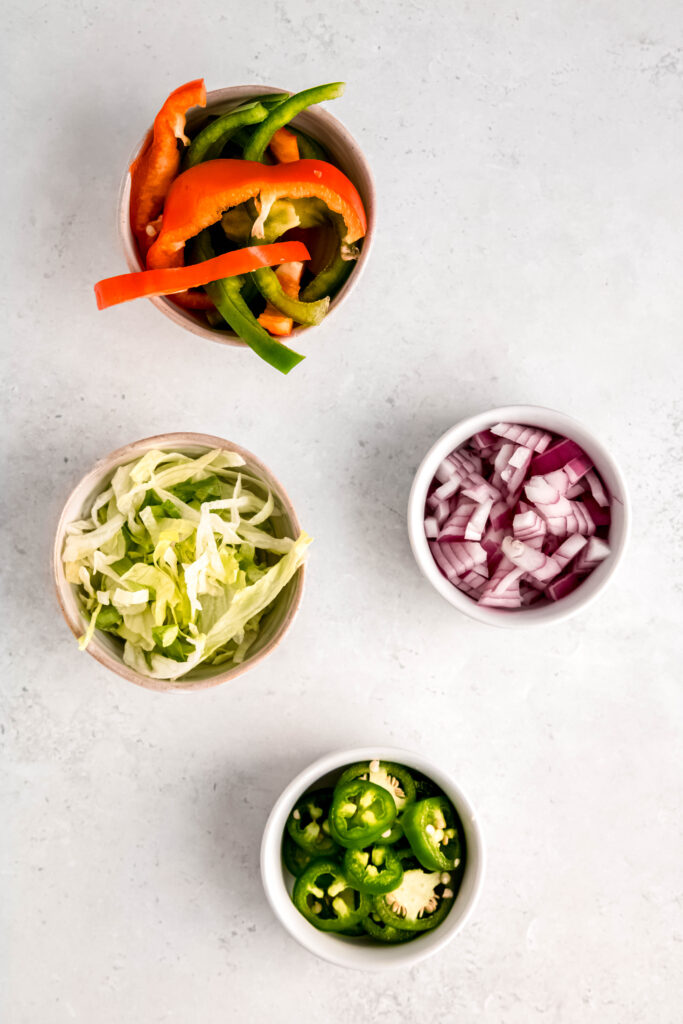 Toppings of your choice in bowls: sliced peppers, shredded lettuce, diced red onion, and chopped jalapeños. 