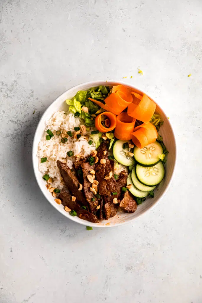 White rice, steak, broccoli, lettuce, carrots, sliced cucumbers in white bowl topped with peanuts and green onion.