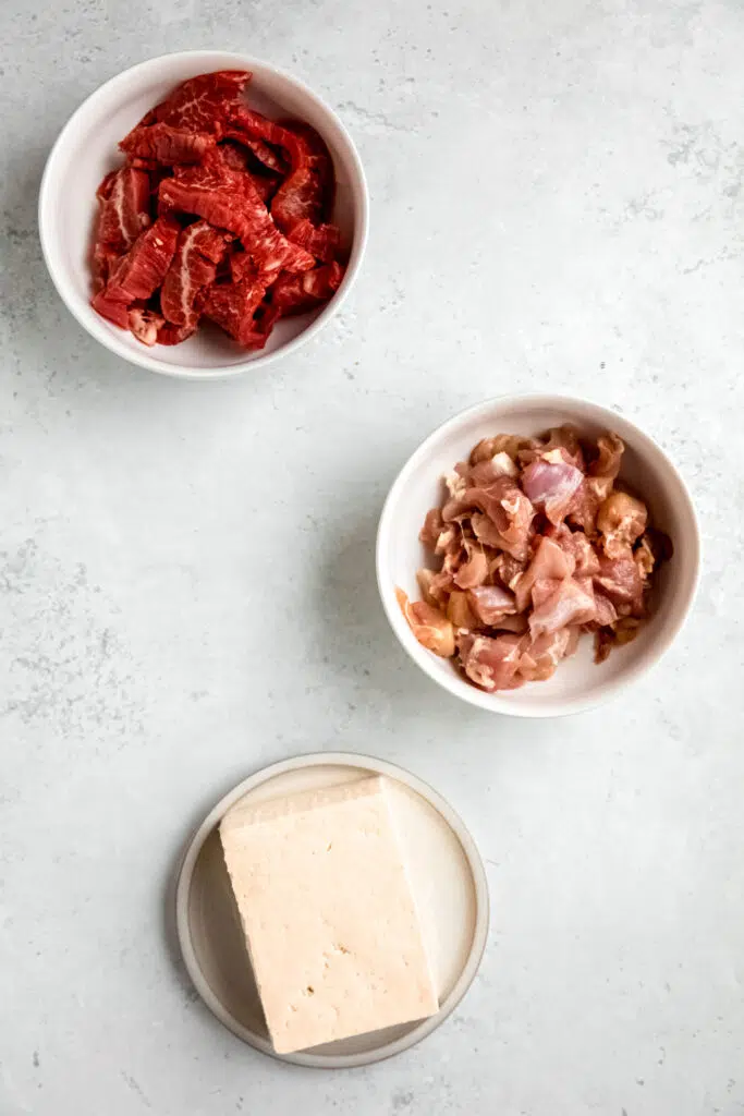 Protein sources in white bowls: raw steak, raw chicken, and tofu.