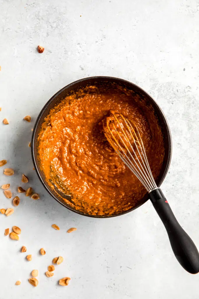 Peanut sauce in black bowl and with a whisk and whole peanuts in background