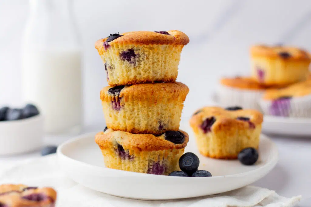 3 blueberry protein muffins stacked on top of each other with fresh blueberries and milk in background.