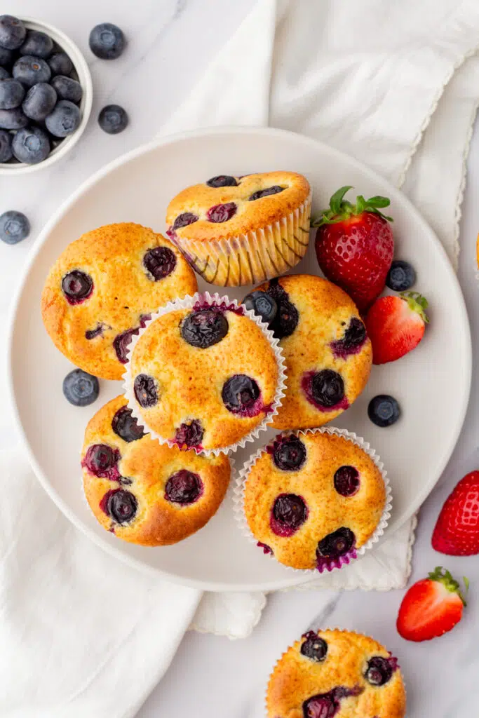 Blueberry protein muffins stacked on on white plate with fresh strawberries and blueberries around.