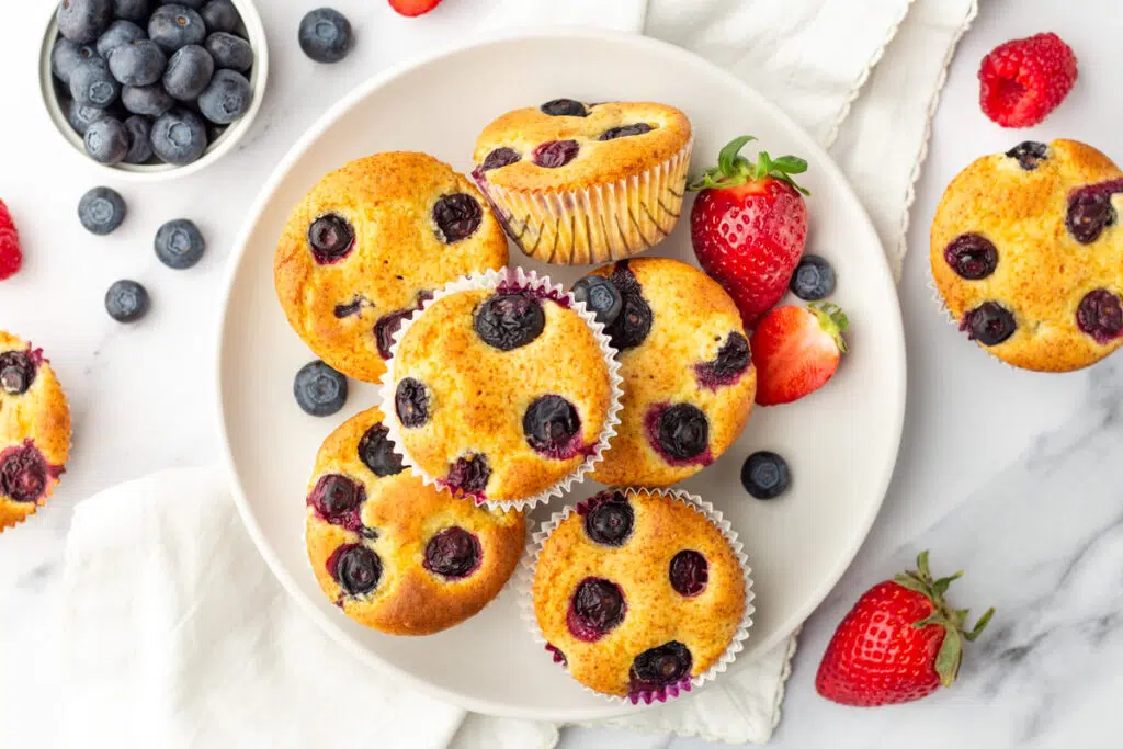 Blueberry protein muffins stacked on white plate with fresh strawberries, blueberries around.