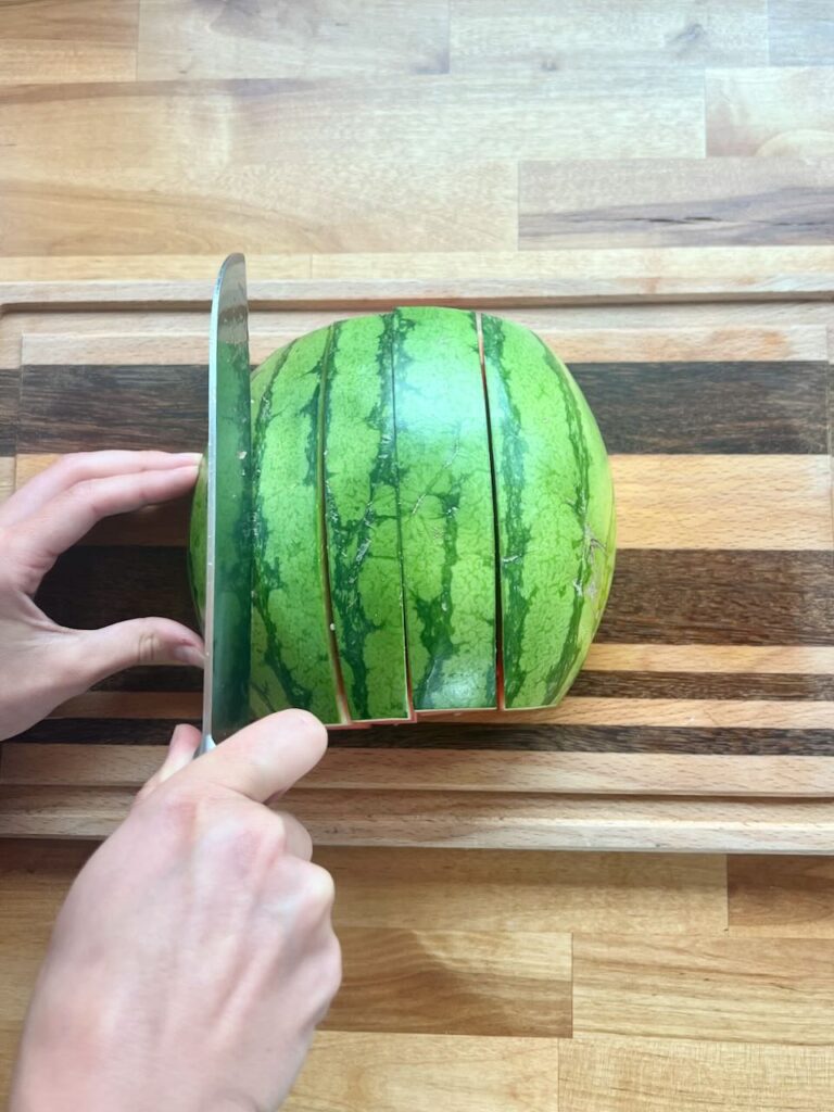 Half of watermelon being cut lengthwise in to even 1-2 inch strips, with rind. 