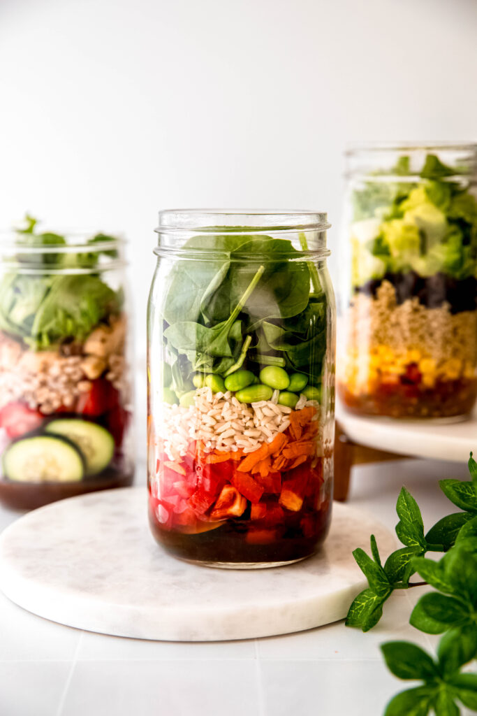 Mason jar with leafy greens on top of edamame, grains, veggies, and dressing