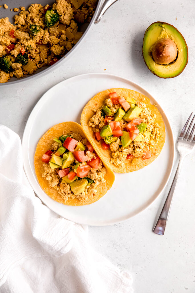 Tofu scramble in corn tortillas topped with avocado and tomatoes on white plate
