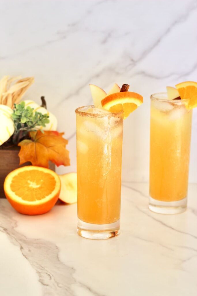 Two apple cider mocktails with orange half and fall decor in background