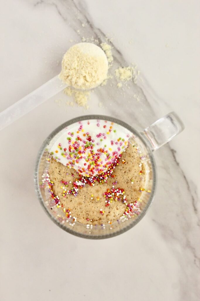 Birthday cake vanilla protein mug cake topped with rainbow sprinkles with protein powder in background