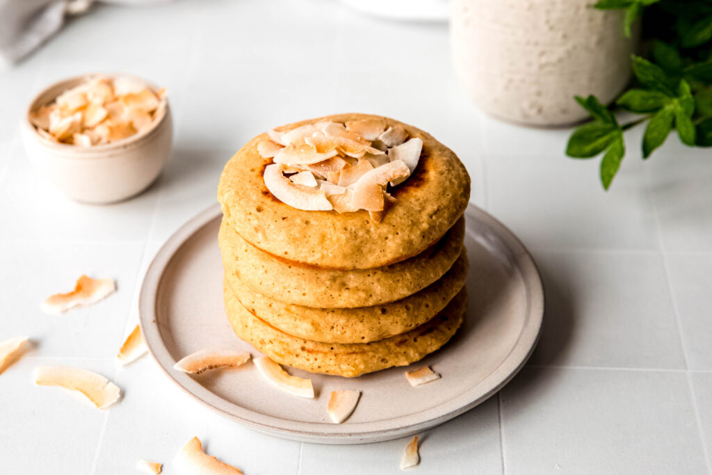 Four protein pancakes stacked topped with shredded coconut on beige plate