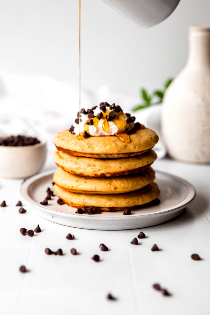 Four protein pancakes stacked topped with whipped cream and chocolate chips, with syrup drizzling on