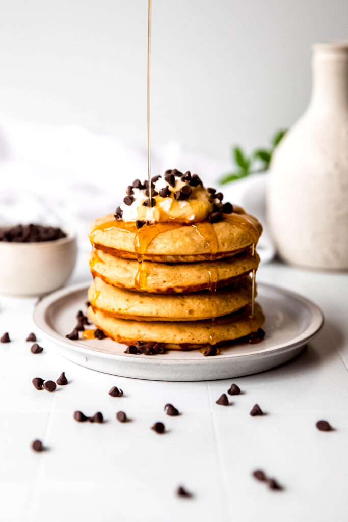 Four protein pancakes stacked  topped with whipped cream and chocolate chips, with syrup drizzling on