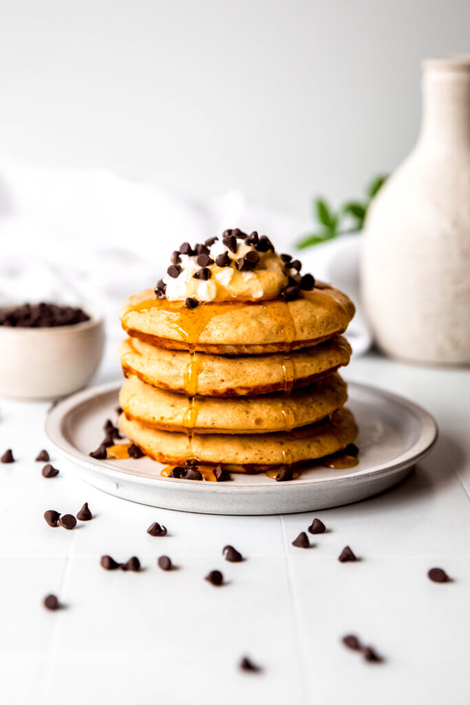 Four protein pancakes stacked  topped with whipped cream and chocolate chips and syrup.