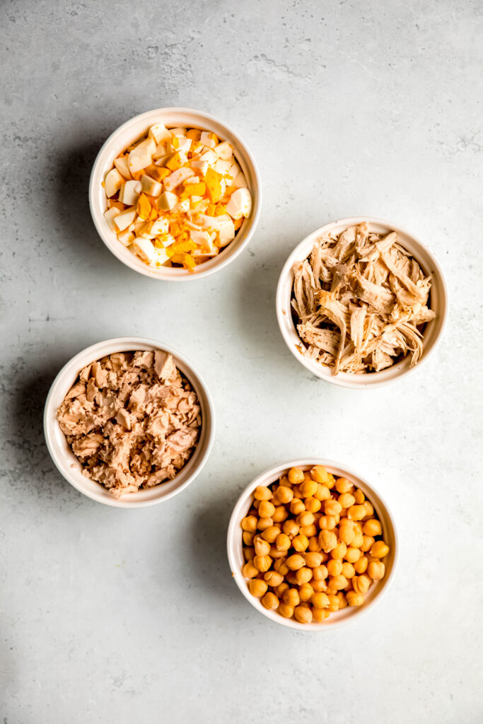Proteins of choice in white bowls: chicken, tuna, chickpeas, hard boiled eggs.